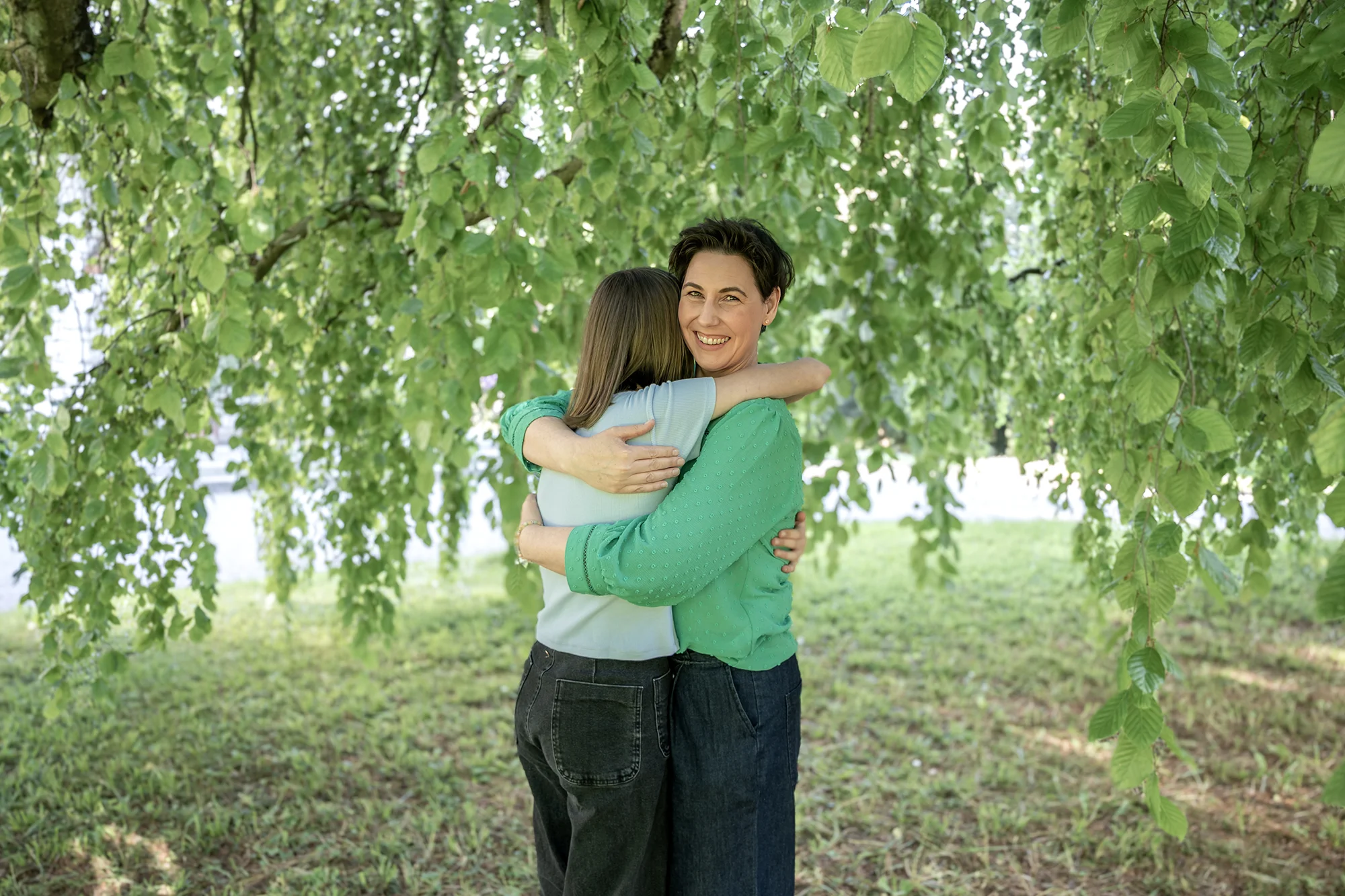 Eine Frau umarmt lächelnd ein Kind unter einem grünen Baum, symbolisiert Unterstützung und die innere Stärke entwickeln.
